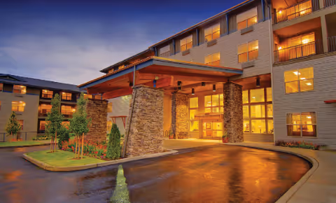 Exterior view of a multi-story senior living facility at dusk with warm lights glowing from the windows. The entrance features a covered drop-off area supported by stone pillars, surrounded by landscaped greenery and a curved driveway.