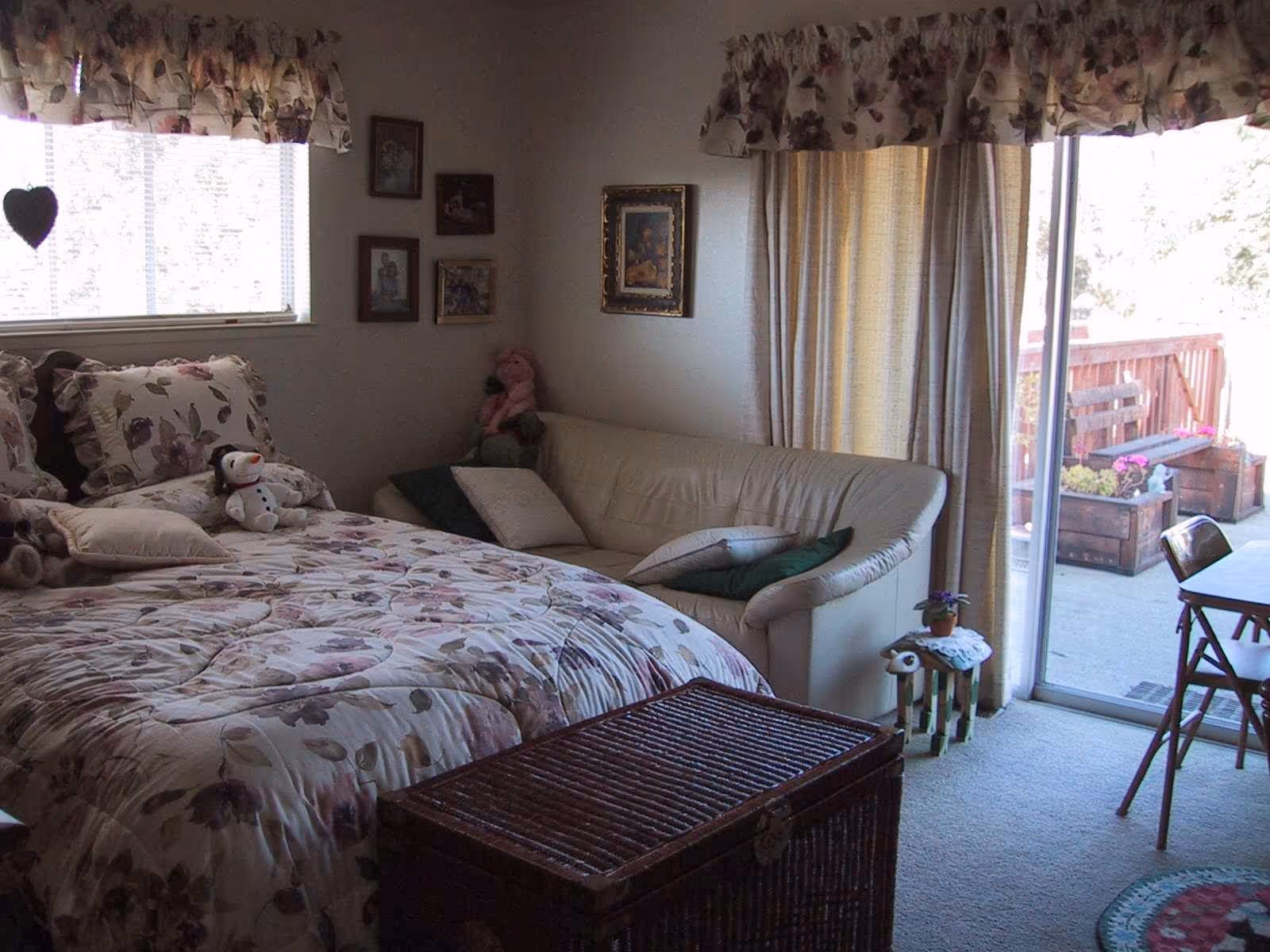 Sunlit bedroom with a floral bedspread, a small loveseat, wicker chest, and sliding glass door opening to an outdoor patio.