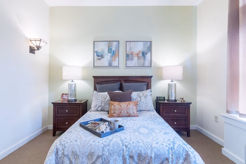 A neatly made bed with patterned bedding and multiple pillows in a bedroom. The bed has a dark wooden headboard and is flanked by two matching dark wooden nightstands, each with a lamp. Above the bed are two abstract paintings. A tray with a cup, glasses, and a book is placed on the bed. The room has light-colored walls and a window with curtains on the right side.