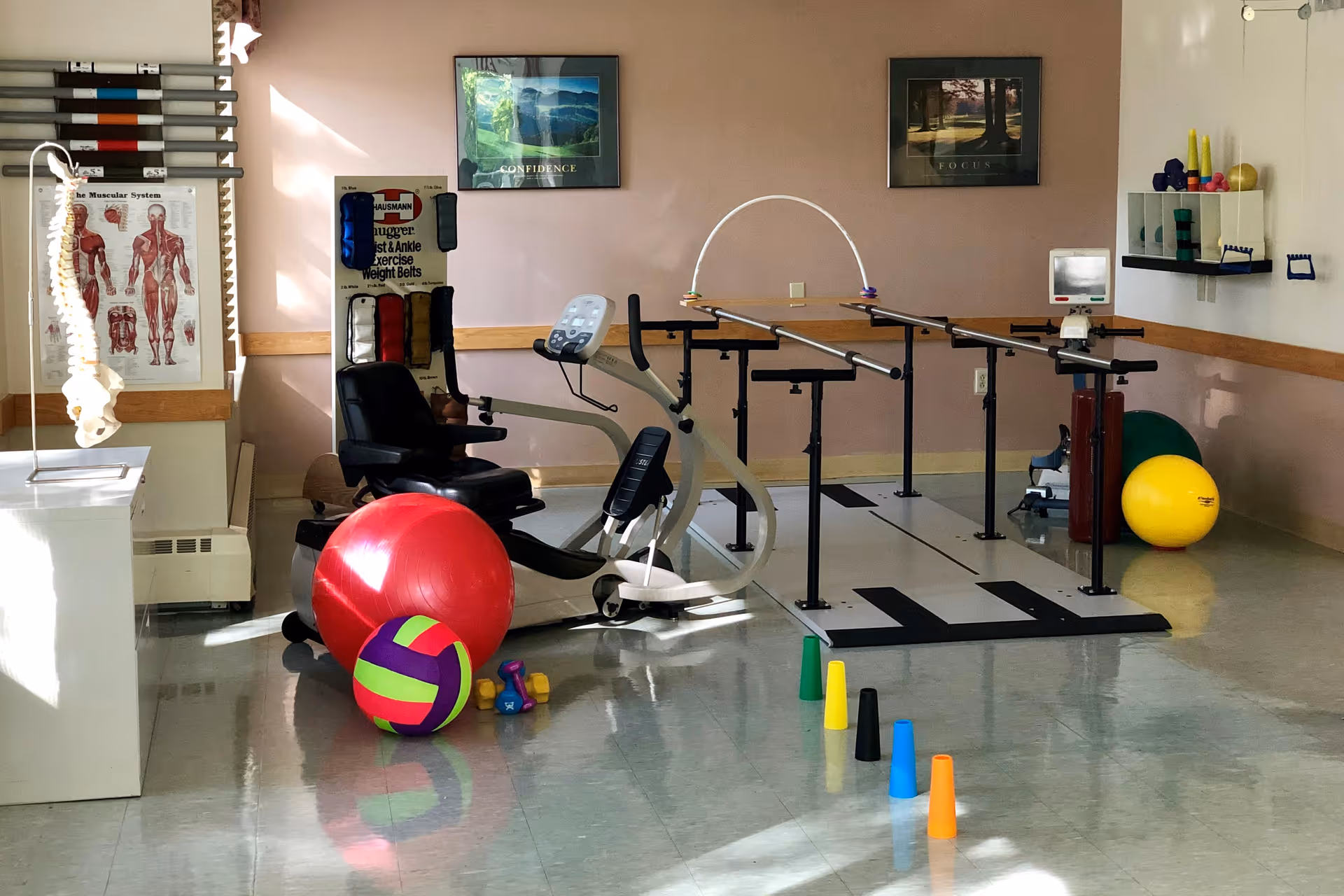 Interior therapy room with parallel bars, an exercise bike, colorful therapy balls and cones.