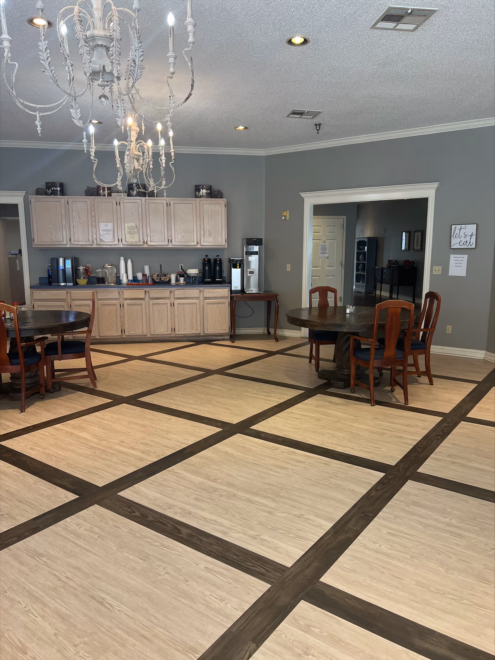 A dining area in a senior living facility with round wooden tables and chairs. There is a chandelier hanging from the ceiling. Along one wall, there are light wood cabinets and a counter with coffee machines, cups, and other beverage supplies. The walls are painted gray, and there is a sign on the wall that says 'let's eat'.