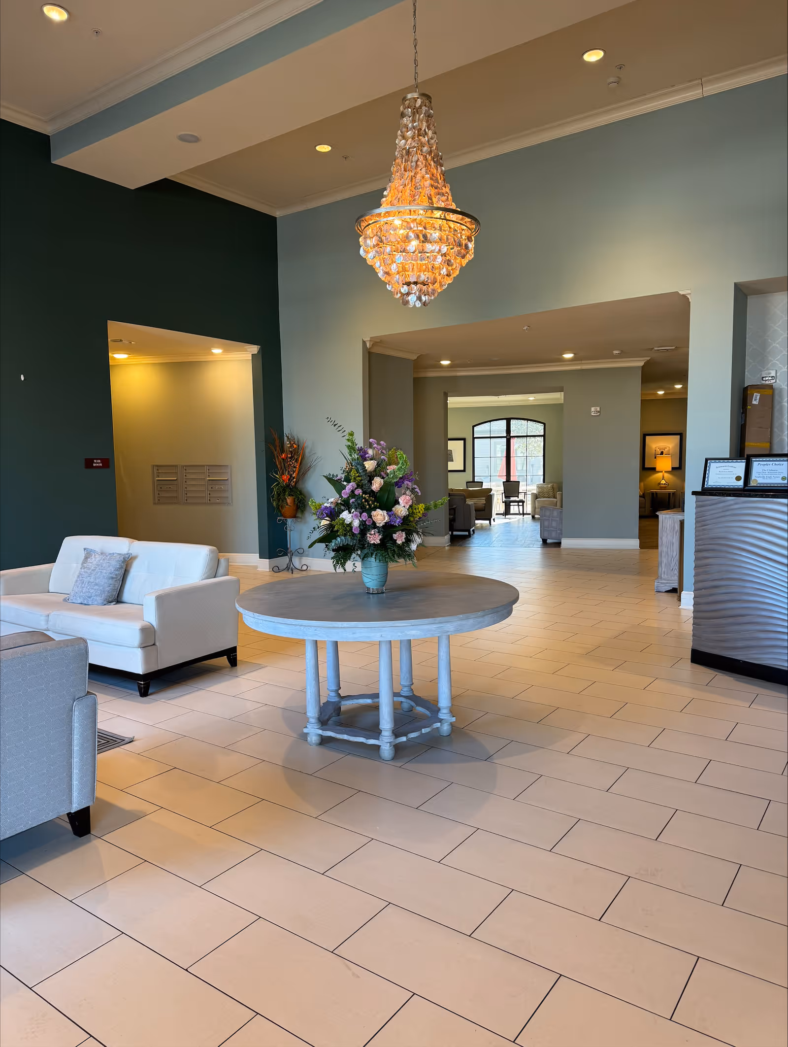 A spacious and well-lit lobby area with a round wooden table in the center holding a large floral arrangement. There is a crystal chandelier hanging above the table. The room features light-colored tiled flooring, a white sofa with a cushion, and a dark green accent wall. In the background, there are more seating areas with armchairs and large windows letting in natural light.