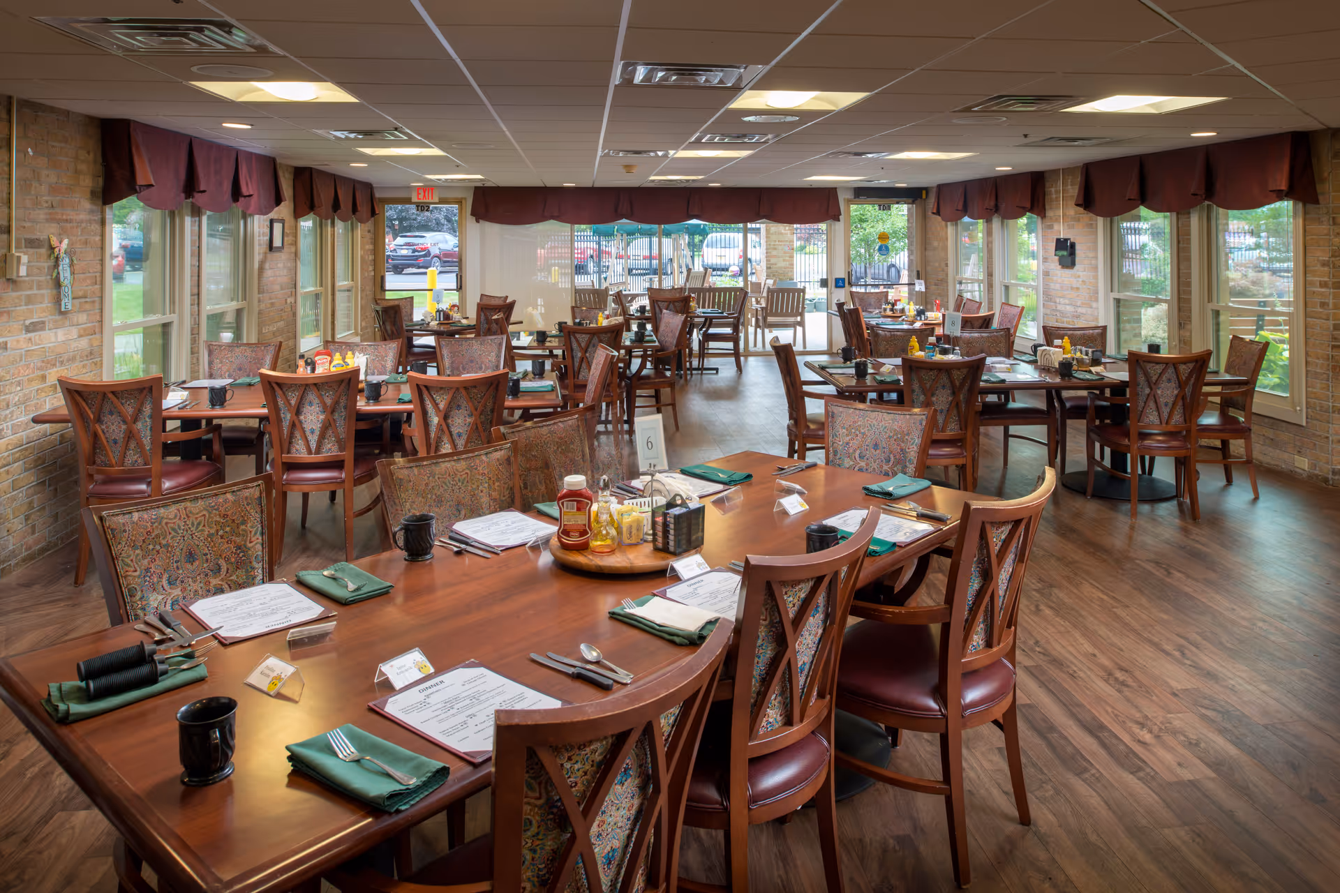Communal dining room with multiple wooden tables and chairs set for meals, large windows, and hardwood floors.