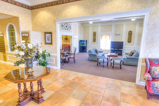 A bright and spacious senior living facility common area with tiled flooring in the foreground and a carpeted seating area in the background. The seating area includes sofas, armchairs, a coffee table, and a television. There is a decorative table with a floral arrangement in the foreground and a fish tank visible in the adjoining room. The walls are adorned with patterned wallpaper and framed artwork.