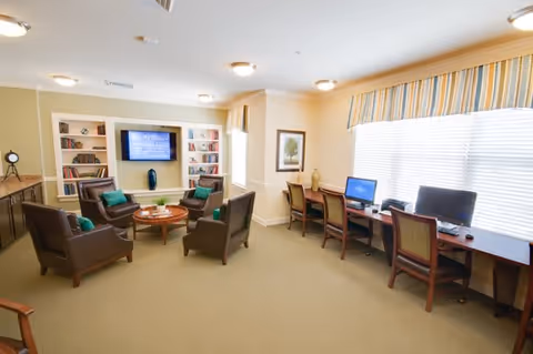 A well-lit common area with a seating arrangement of four brown armchairs around a round wooden coffee table. Behind the seating area is a built-in bookshelf with decorative items and a flat-screen TV mounted in the center. To the right, there is a long desk with three chairs and two desktop computers placed on it, positioned in front of a large window with striped valances. The room has beige walls and carpeted flooring.