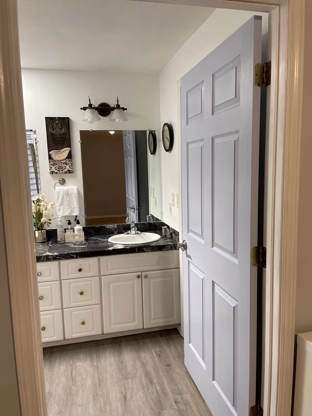 View into a bathroom with a white door partially open, showing a vanity with a black marble countertop, a white sink, a large mirror, and a wall-mounted light fixture above the mirror. There are decorative items including a small plant, soap dispensers, a towel hanging on a ring, and a wall clock.