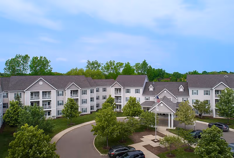 Exterior view of a large, three-story senior living facility building with multiple windows and balconies, surrounded by green trees and a curved driveway with parked cars under a partly cloudy sky.