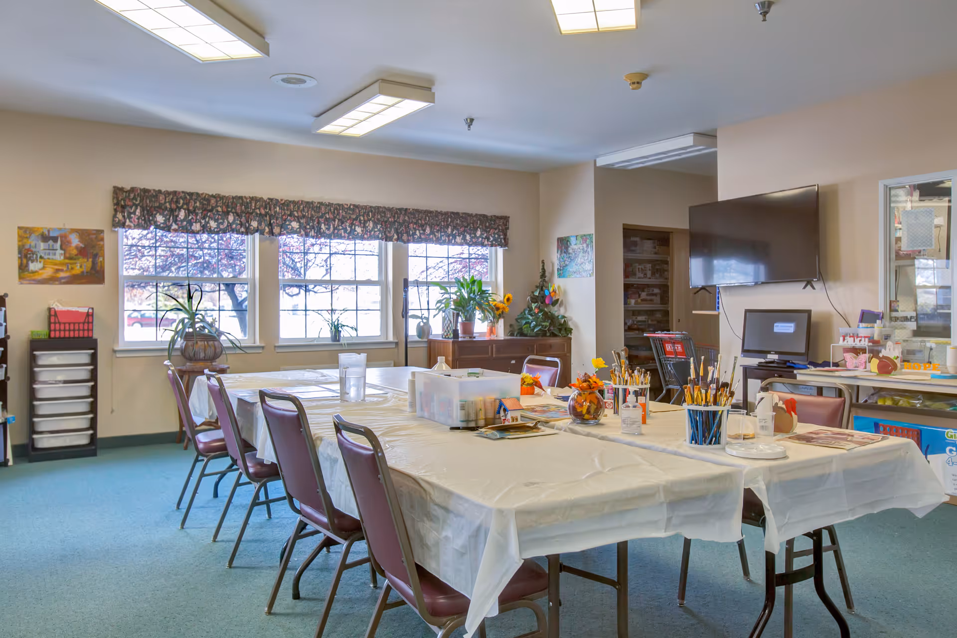 A bright activity room with long tables covered in plastic, chairs, craft supplies, potted plants and a wall-mounted TV.