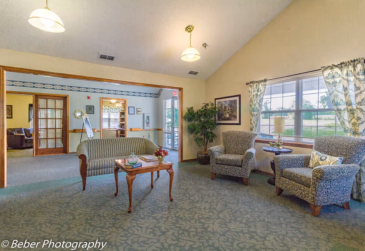 A cozy senior living facility common area with patterned carpet, two upholstered armchairs, a small round table with a lamp, a striped loveseat, and a wooden coffee table with books and a flower vase. Large windows with floral curtains let in natural light. The room opens into another area with additional seating and a glass door leading outside.