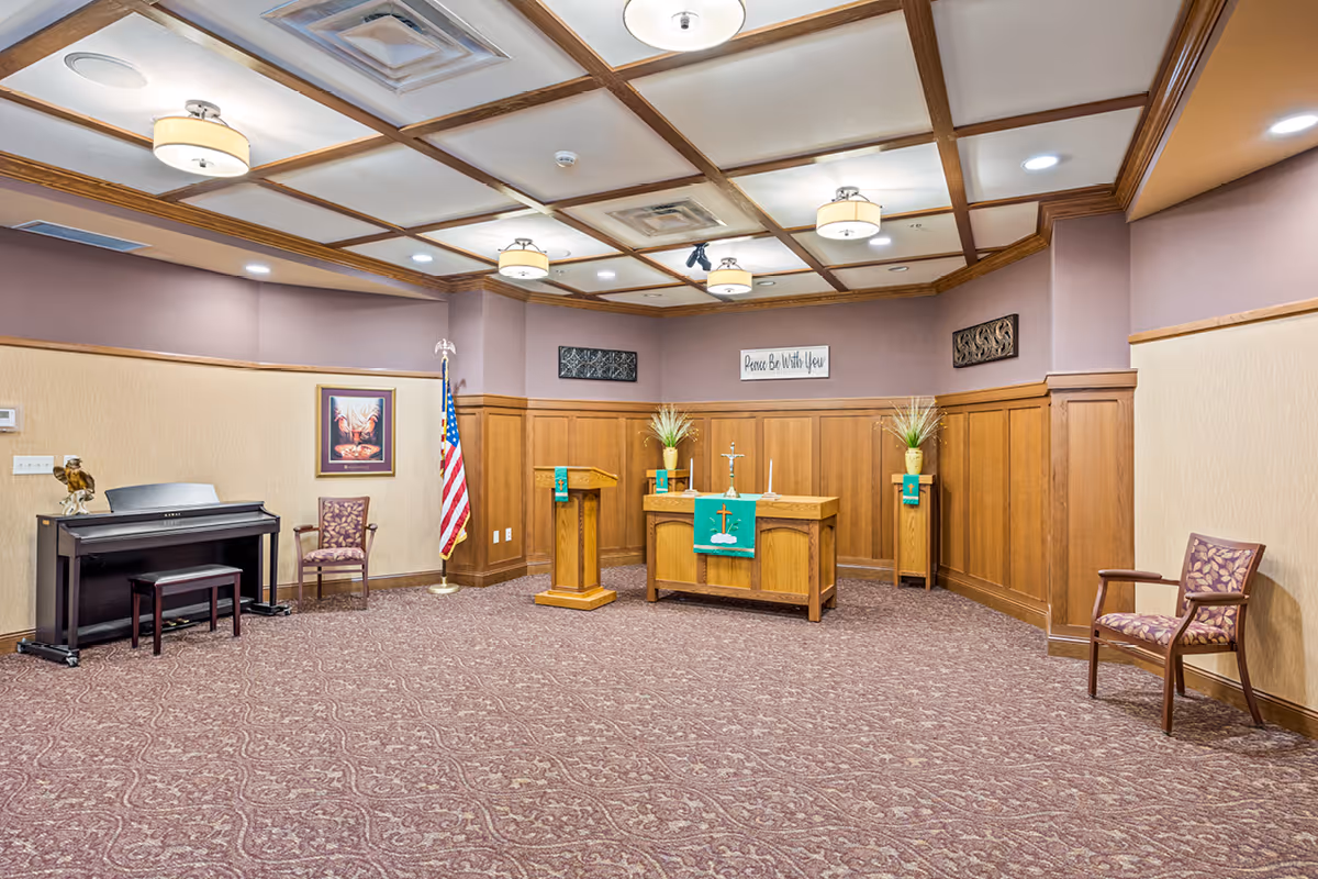 A small chapel or worship room with a wooden altar draped with a green cloth featuring a cross, two candles, and two flower arrangements on stands. There is a wooden podium, an American flag, a piano with a bench, and two upholstered chairs. The walls are decorated with framed artwork and a sign that reads 'Peace Be With You'. The ceiling has wooden beams and multiple light fixtures, and the floor is carpeted with a patterned design.