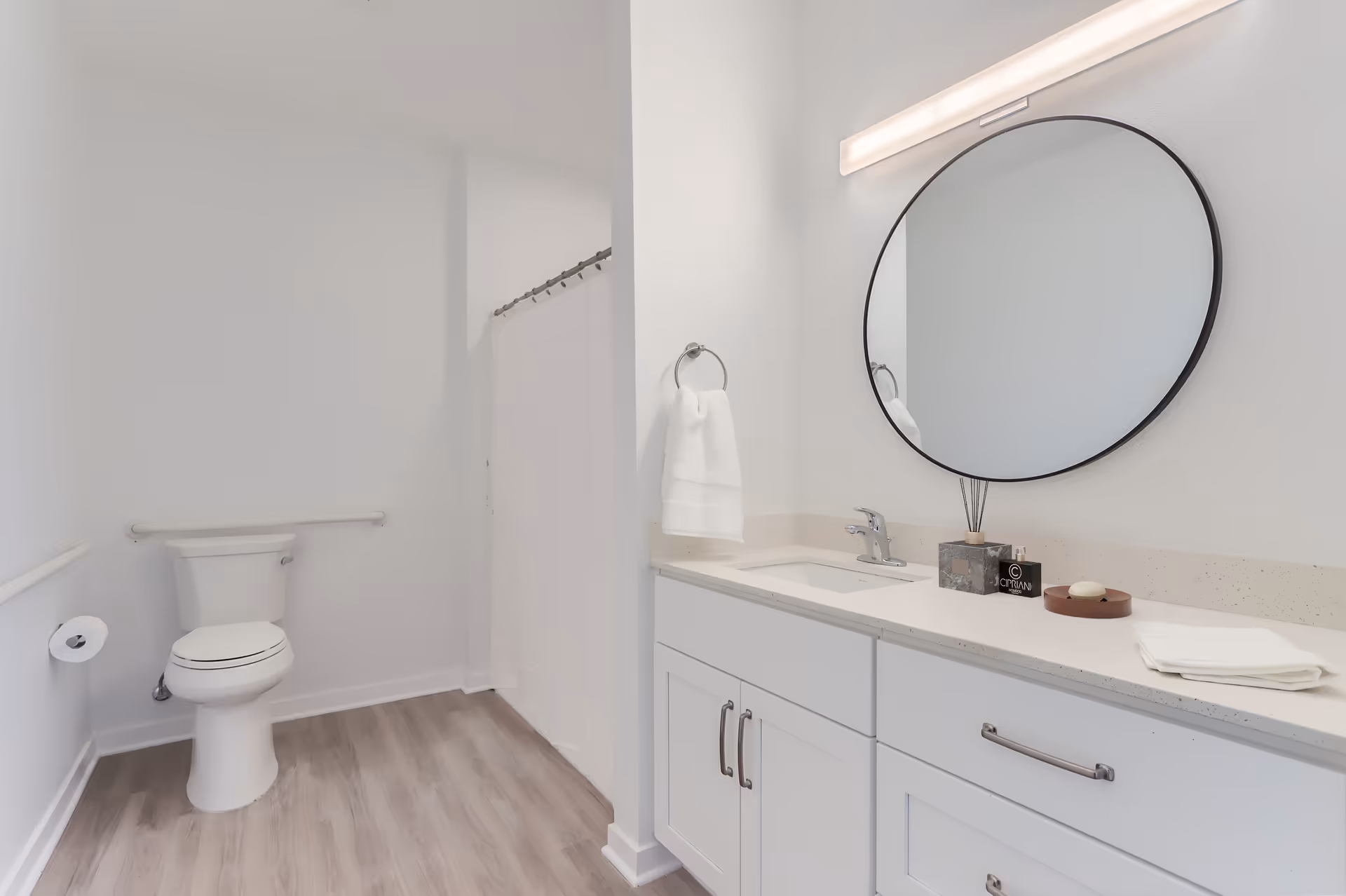 Well-lit modern bathroom with a toilet, a white vanity and sink topped by a round mirror, and a shower area with a curtain.