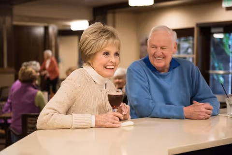Two smiling senior residents sit at a countertop in a communal dining/lounge area, one holding a glass of red wine.