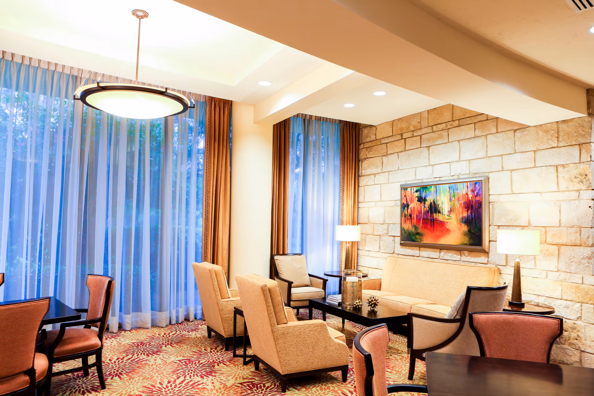 A cozy and well-lit living room area with beige armchairs and a sofa arranged around a dark coffee table. The room features a stone accent wall with a colorful abstract painting, two table lamps, large windows with sheer and brown curtains, and a patterned carpet with red and beige tones. There are also dining tables and chairs visible in the foreground.