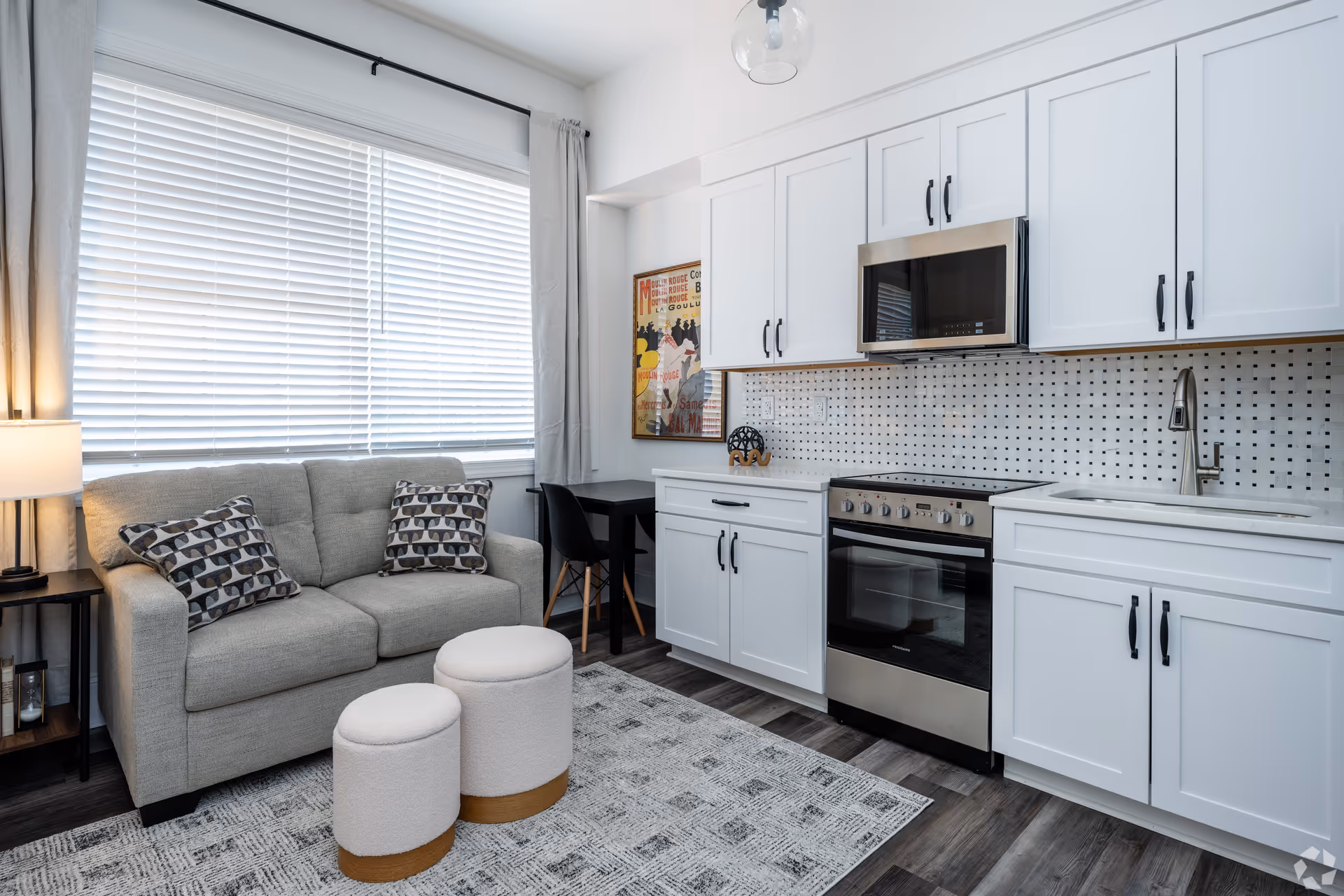 A modern apartment interior featuring a small kitchen area with white cabinets, a stainless steel oven and microwave, and a sink. Adjacent to the kitchen is a cozy living space with a beige loveseat, two patterned pillows, two round ottomans, a side table with a lamp, and a window with white blinds and light curtains. A small black table with a chair is positioned in the corner near a colorful wall art piece.