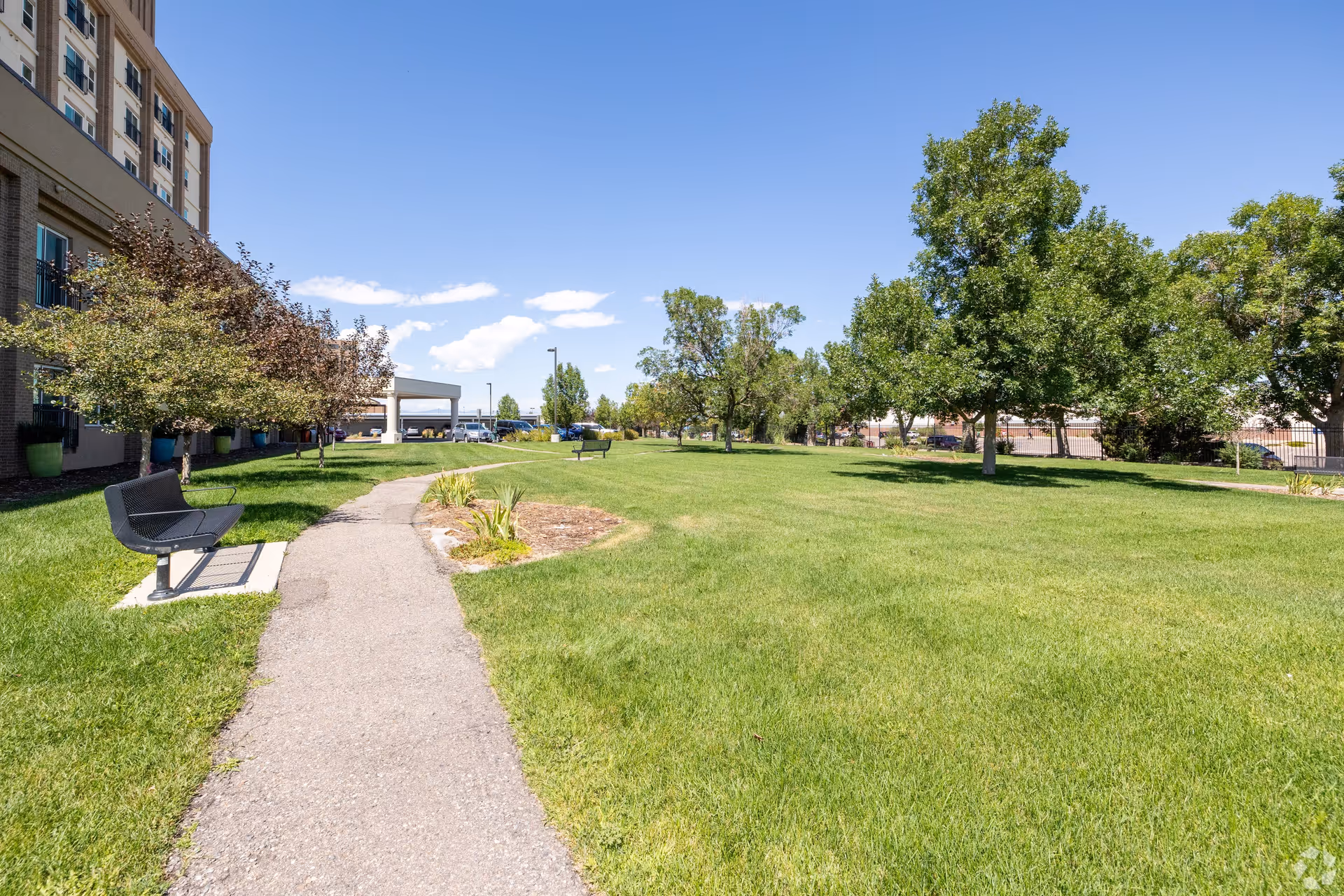 A sunny outdoor area at Bella Vita featuring a paved walking path, green grass, several trees, and benches along the path. The side of a multi-story building is visible on the left with a clear blue sky overhead.