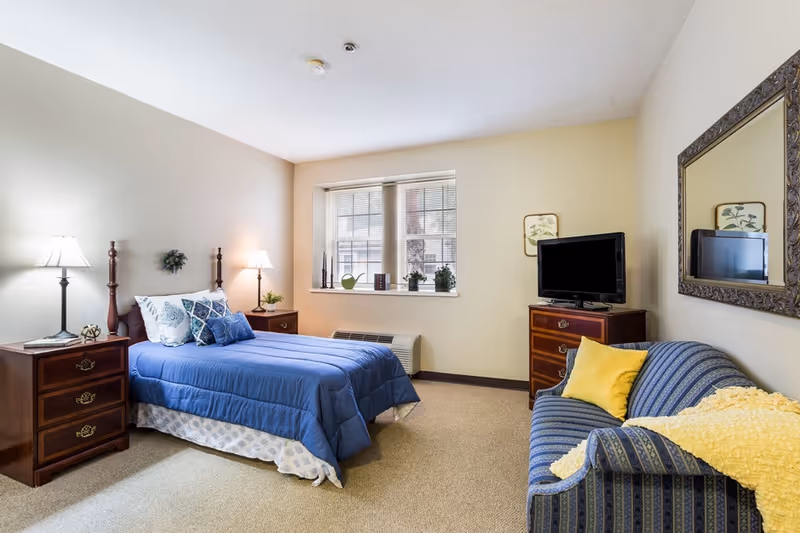 A cozy bedroom in an assisted living facility featuring a bed with blue bedding and multiple pillows, two wooden nightstands with lamps, a window with blinds, a wooden dresser with a flat-screen TV on top, a blue patterned couch with a yellow pillow and yellow throw blanket, and a large decorative mirror on the wall.