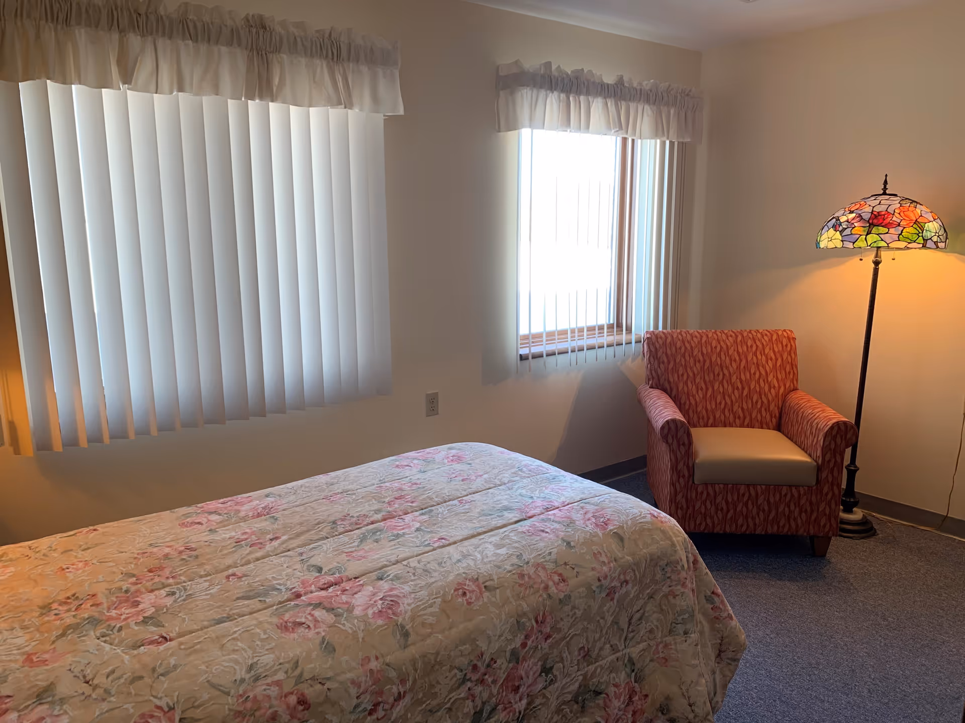 A small bedroom with a floral bedspread, an upholstered armchair, a stained-glass floor lamp, and windows with vertical blinds.