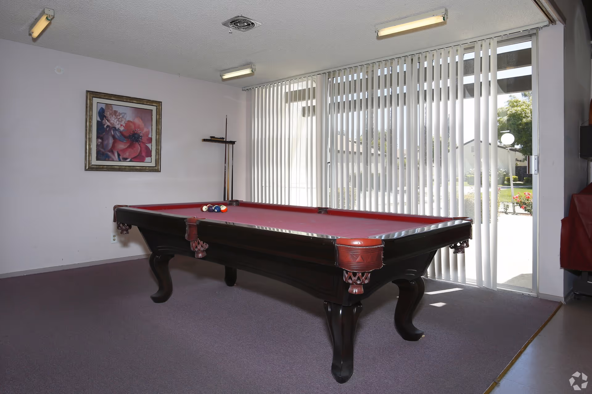 A red-felt pool table in a carpeted indoor recreation room with vertical blinds, a floral painting, and pool cues.
