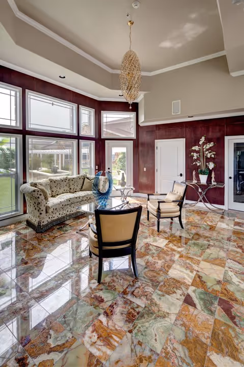 A bright and elegant living room with large windows allowing natural light to fill the space. The room features a patterned sofa, two beige armchairs, a glass coffee table, and a side table with white flowers. The floor is covered with colorful marble tiles, and a decorative chandelier hangs from the ceiling.