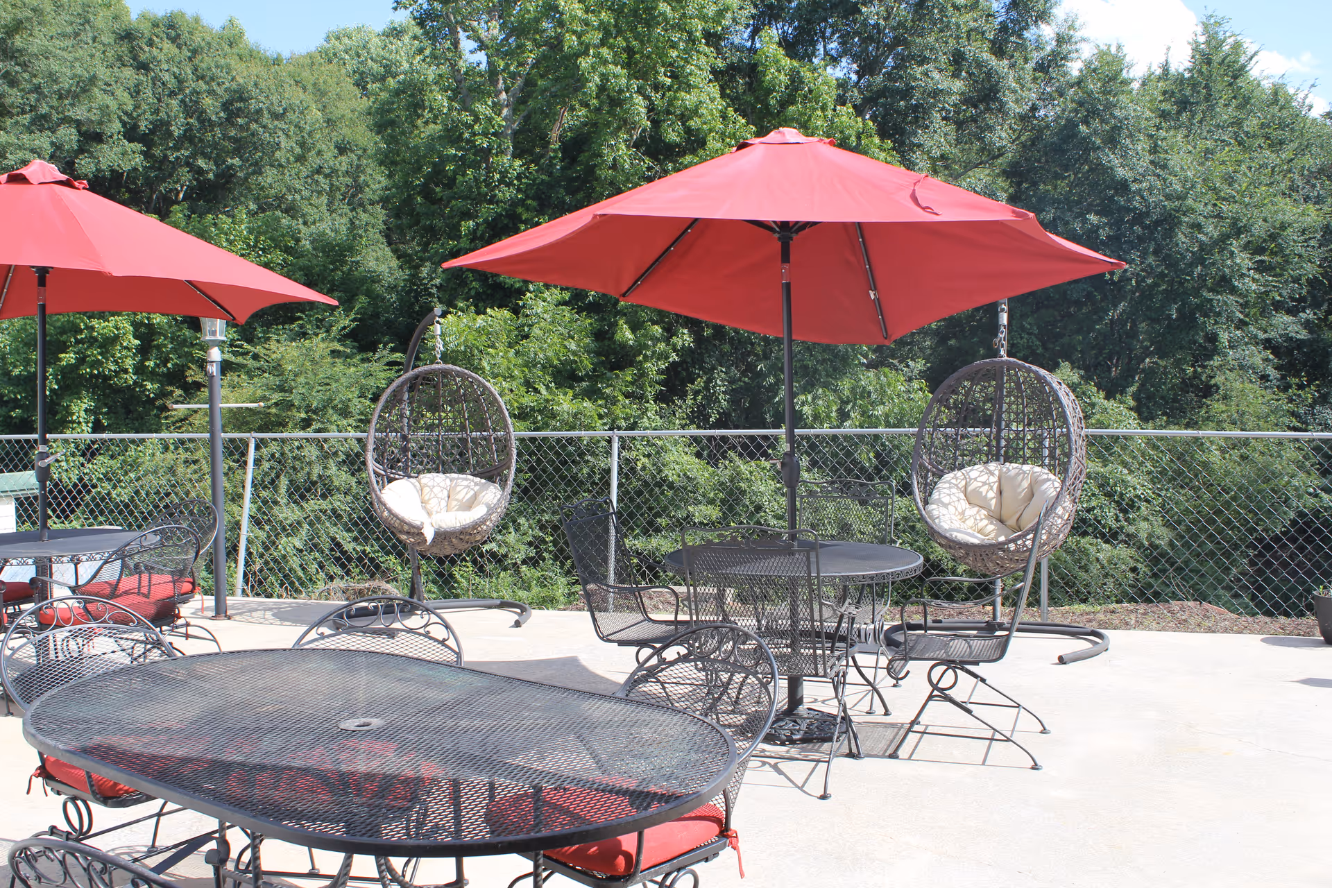 Outdoor patio area with black metal tables and chairs featuring red cushions, two red umbrellas providing shade, and two hanging wicker chairs with white cushions. The patio is surrounded by a chain-link fence and lush green trees in the background under a clear blue sky.
