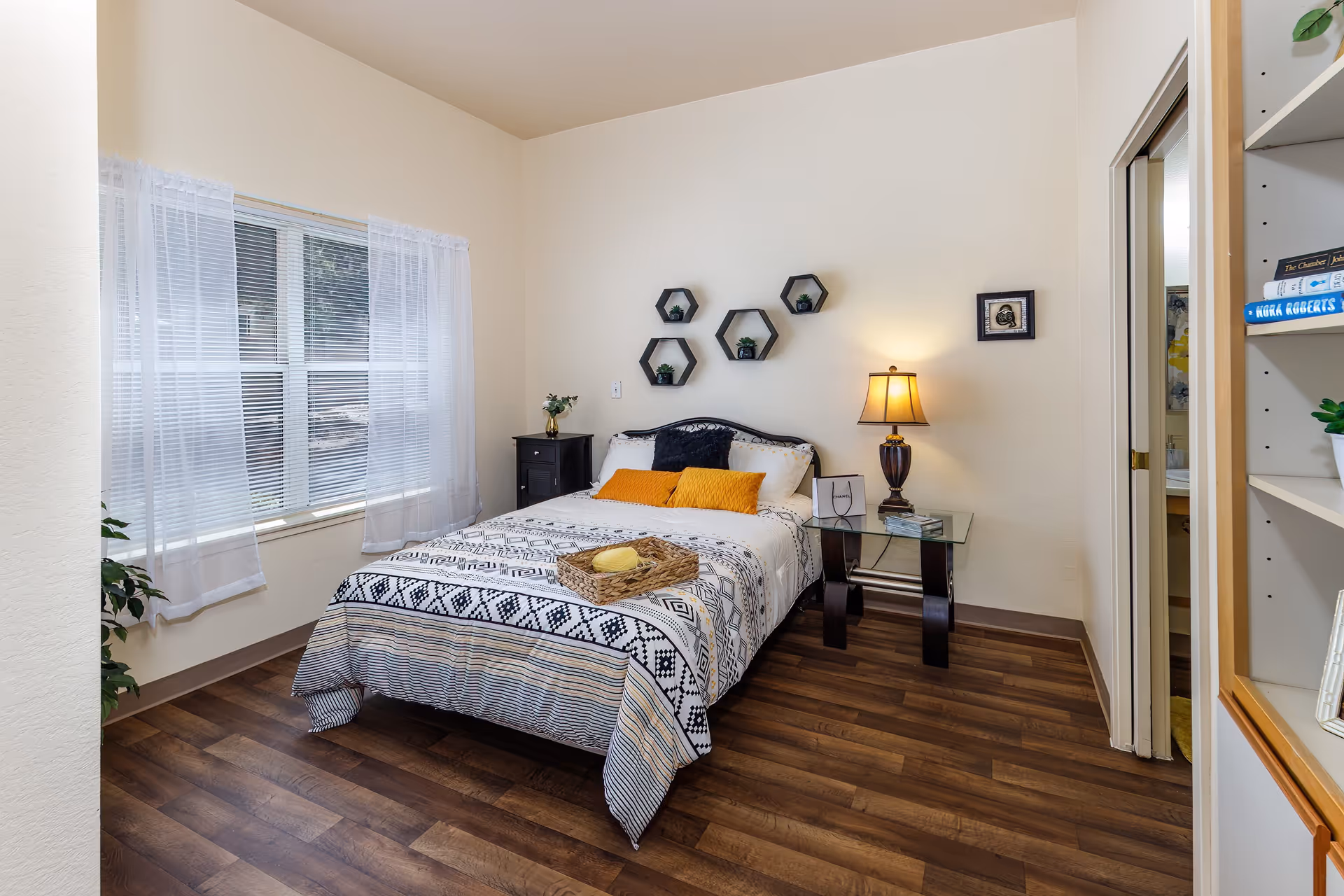 A cozy bedroom with a double bed covered in a patterned bedspread and two yellow pillows. The room features a wooden floor, a window with sheer white curtains, a black nightstand with a small plant, a glass side table with a lamp, and hexagonal wall shelves with small plants. A doorway leads to a bathroom, and a bookshelf is partially visible on the right.