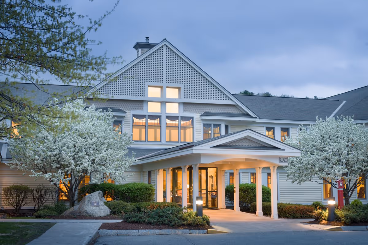Exterior view of a senior living facility named Harvest Hill during dusk, showing a large building with white siding, a peaked roof, and a covered entrance. The entrance is illuminated with warm lights, and there are blooming white flowering trees and well-maintained shrubs around the walkway leading to the door.