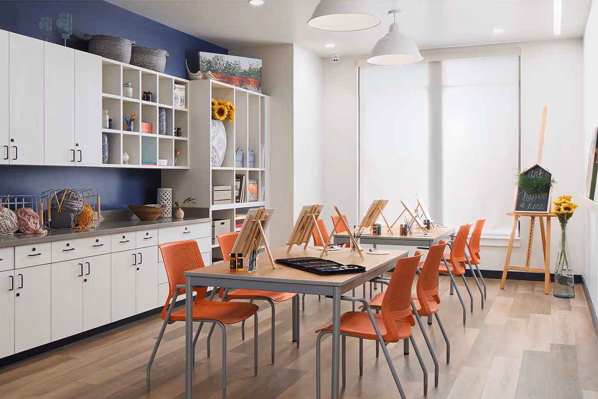 A bright art room with two tables arranged in a row, each with orange chairs and wooden easels holding blank canvases. The room has light wood flooring, white walls, and large windows covered with white blinds. On the left side, there are white cabinets and open shelves filled with art supplies and decorative items. A small chalkboard on an easel near the window reads 'Art happens here,' and a vase with sunflowers is placed nearby.