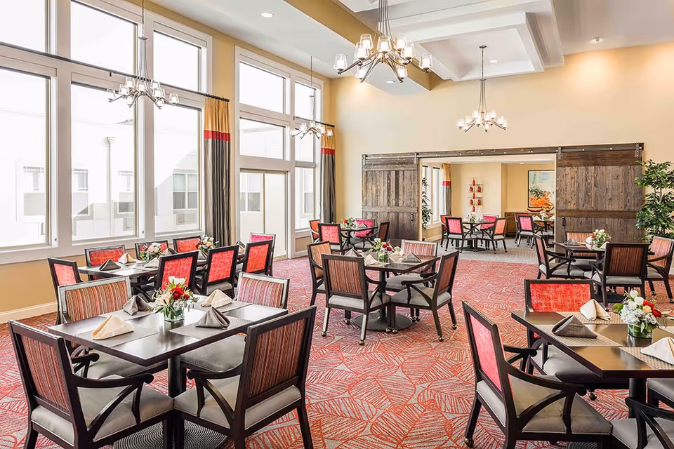 A bright and spacious dining room with multiple tables and chairs arranged neatly. Each table is set with folded napkins and small floral centerpieces. Large windows allow natural light to fill the room, and there are modern chandeliers hanging from the ceiling. The room features a patterned red carpet and wooden sliding barn doors leading to another dining area.