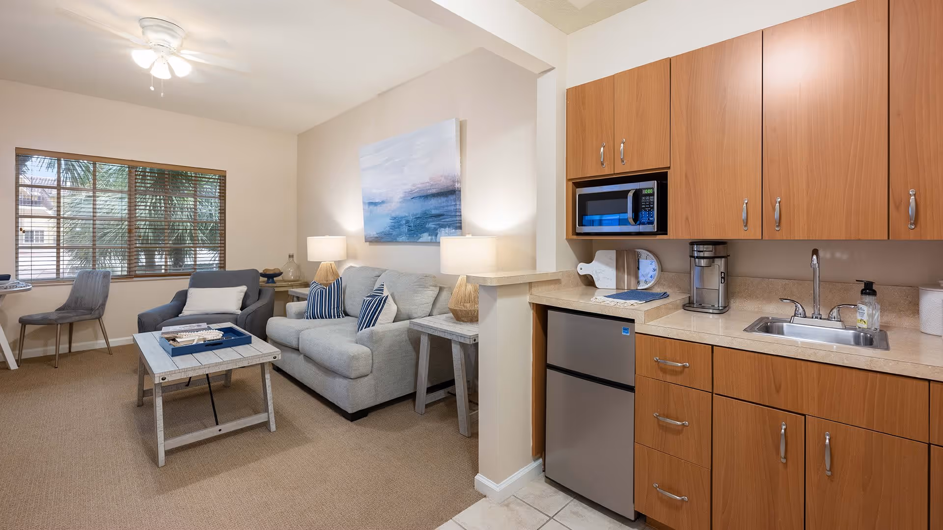 A cozy living area with a gray couch, two blue and white striped pillows, a gray armchair, and a wooden coffee table with a tray on it. There are two side tables with lamps on either side of the couch. A large window with wooden blinds lets in natural light. Adjacent to the living area is a small kitchenette with wooden cabinets, a microwave, a mini refrigerator, a coffee maker, a sink, and a soap dispenser. The room has beige carpet and tiled flooring in the kitchenette area.