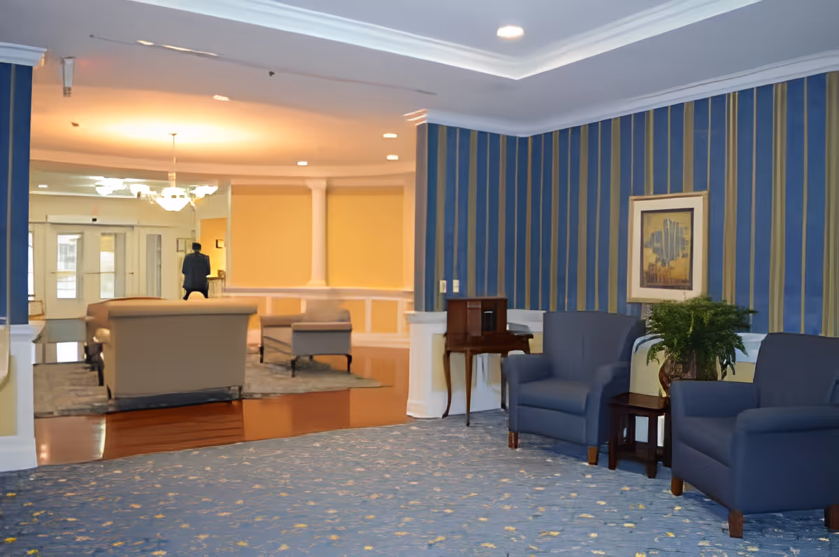 A spacious and well-lit assisted living community common area with blue and gold striped wallpaper, two blue armchairs, a small wooden table with a plant, a framed picture on the wall, and beige sofas in the background near the entrance doors.