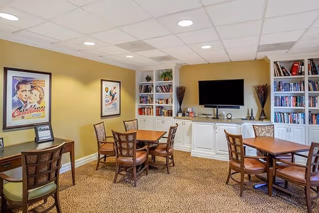 A cozy common area with multiple wooden tables and chairs arranged on a patterned carpet. The walls are painted yellow and decorated with framed vintage movie posters. Built-in white bookshelves filled with books flank a flat-screen TV mounted on the wall above white cabinetry. The ceiling has recessed lighting.