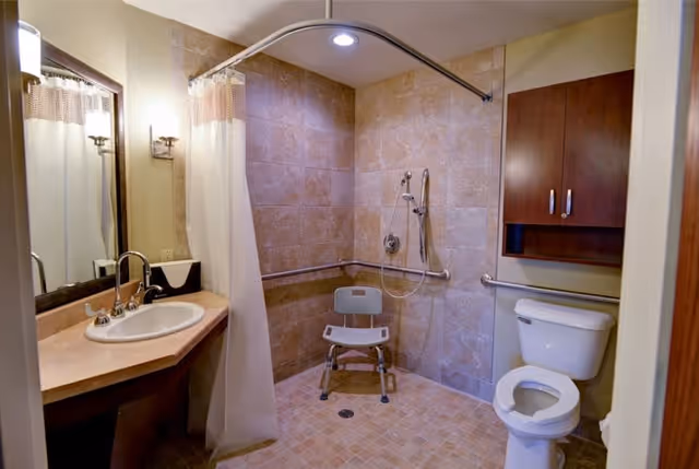 A bathroom with beige tiled walls and floor, featuring a walk-in shower area with a white shower chair and grab bars. There is a white toilet with a wooden cabinet above it, and a sink with a countertop and mirror on the left side. A white shower curtain is partially drawn around the shower area.