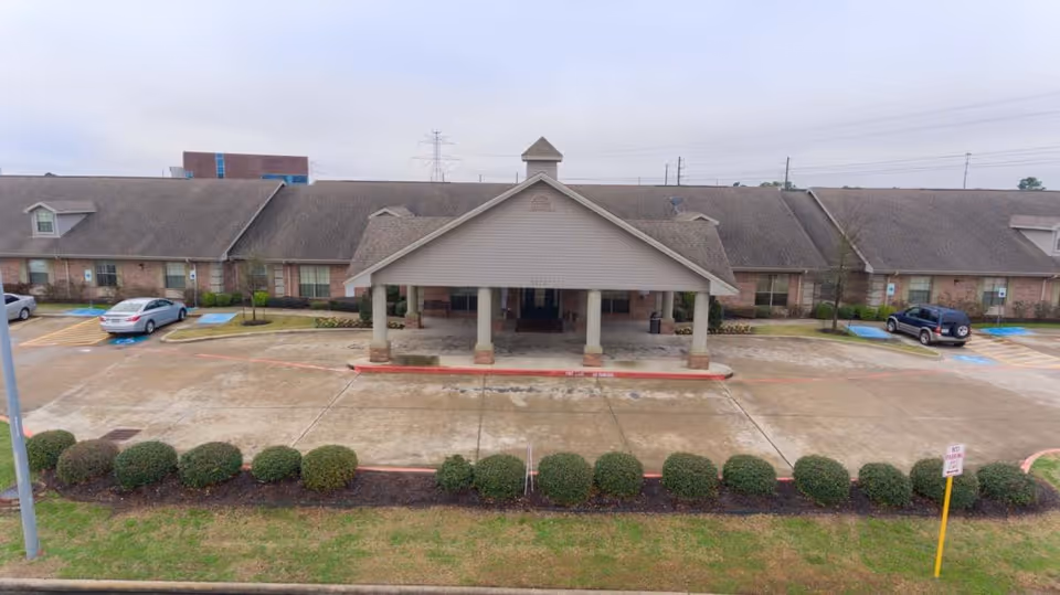 Front exterior view of Mason Creek Transitional Care of Katy, showing a single-story building with a covered entrance supported by columns, a parking area with several cars, and neatly trimmed bushes along the front.