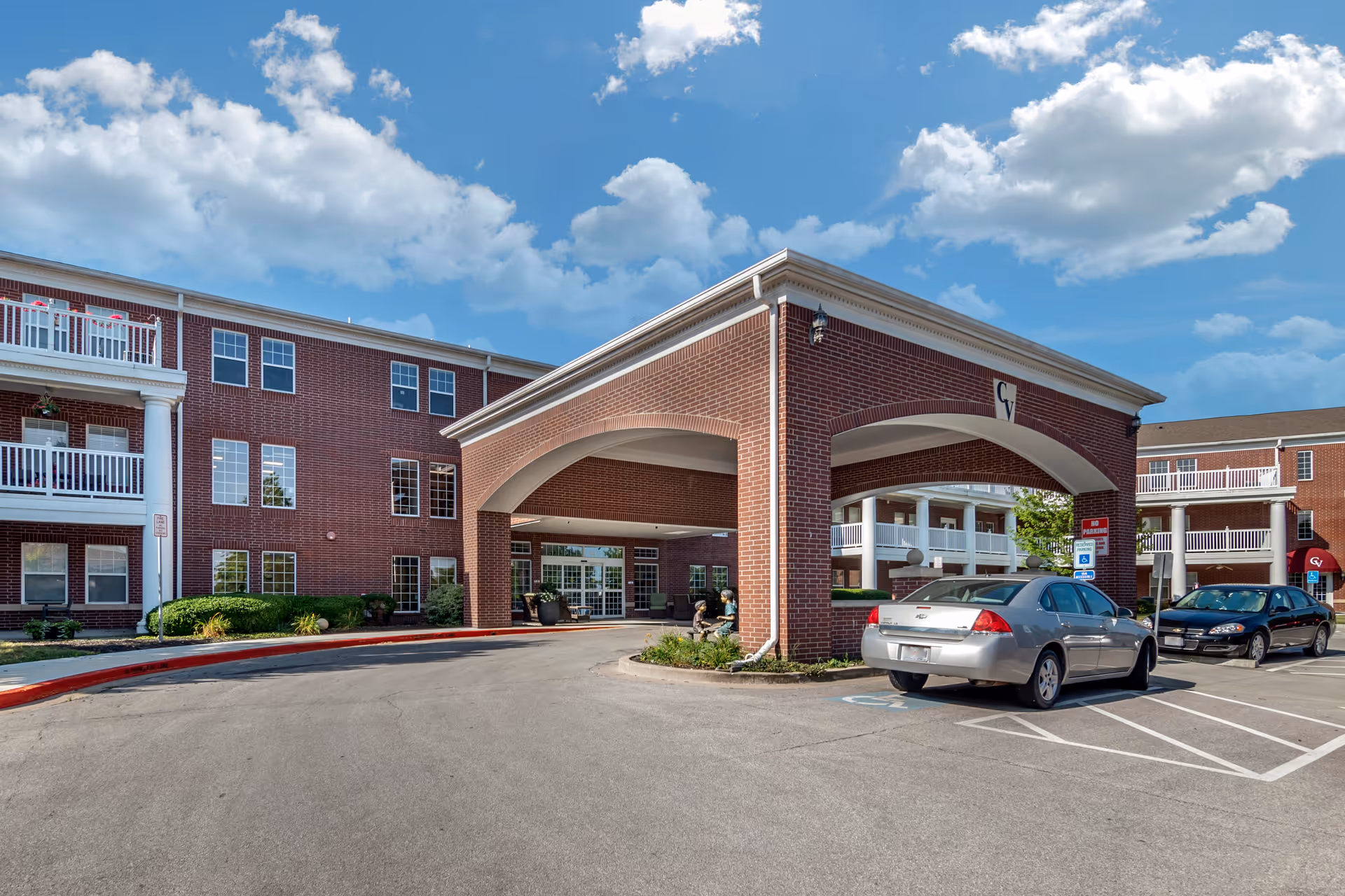 Exterior view of Carnegie Village Senior Living Community showing a brick building with white balconies and a covered entrance with arches. Two cars are parked near the entrance under a partly cloudy blue sky.