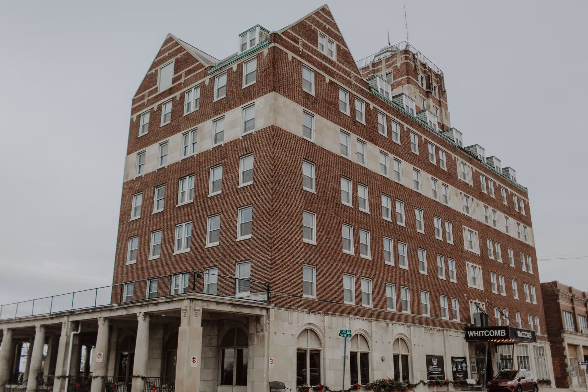 Large multi-story brick senior living tower with arched ground floor and a 'Whitcomb' entrance sign.