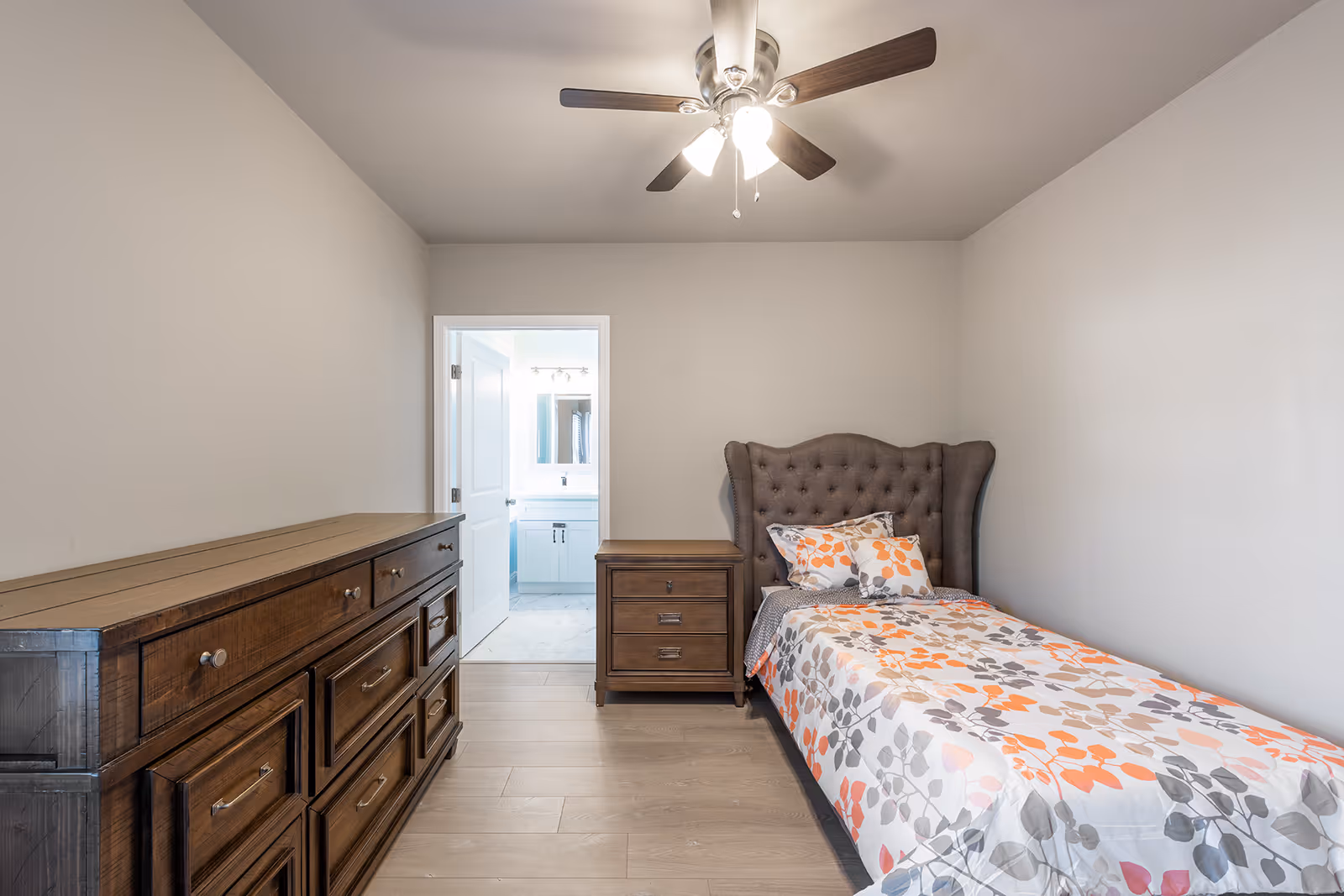 A small bedroom with a single bed featuring a floral patterned bedspread in orange, gray, and white. The bed has a tufted headboard. Next to the bed is a wooden nightstand with two drawers. Across from the bed is a large wooden dresser with multiple drawers. A ceiling fan with lights is mounted on the ceiling. An open door reveals a bathroom with a white vanity and mirror.