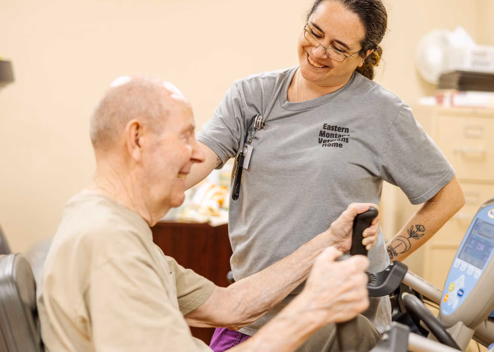 A smiling caregiver wearing a gray shirt with 'Eastern Montana Veterans Home' printed on it assists an elderly man who is using a seated exercise machine indoors.