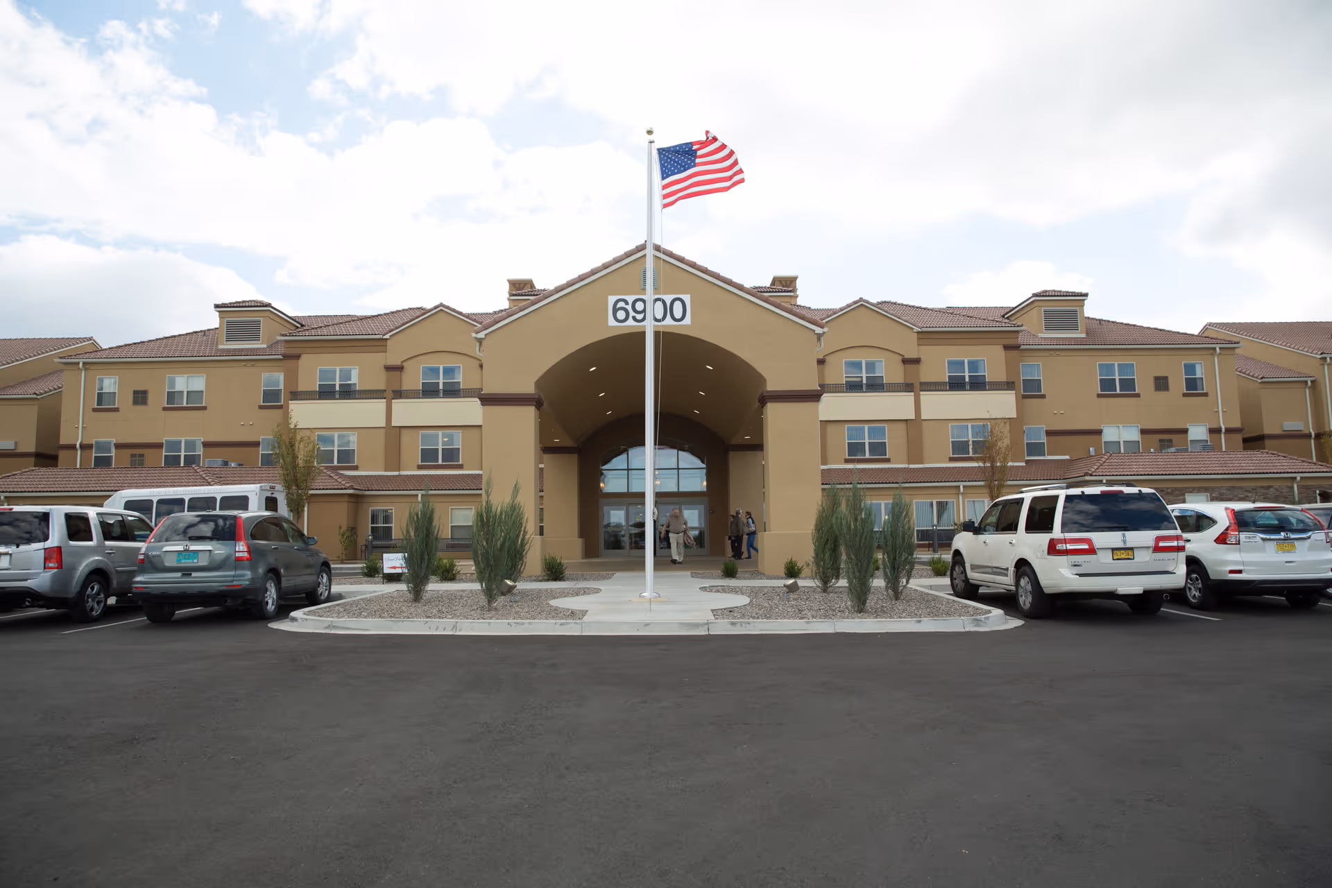 Front exterior view of a large, three-story retirement resort building with a beige facade and red-tiled roof. An American flag is flying on a flagpole in the center of the paved driveway leading to the main entrance. Several cars are parked on either side of the driveway, and a few people are visible near the entrance.