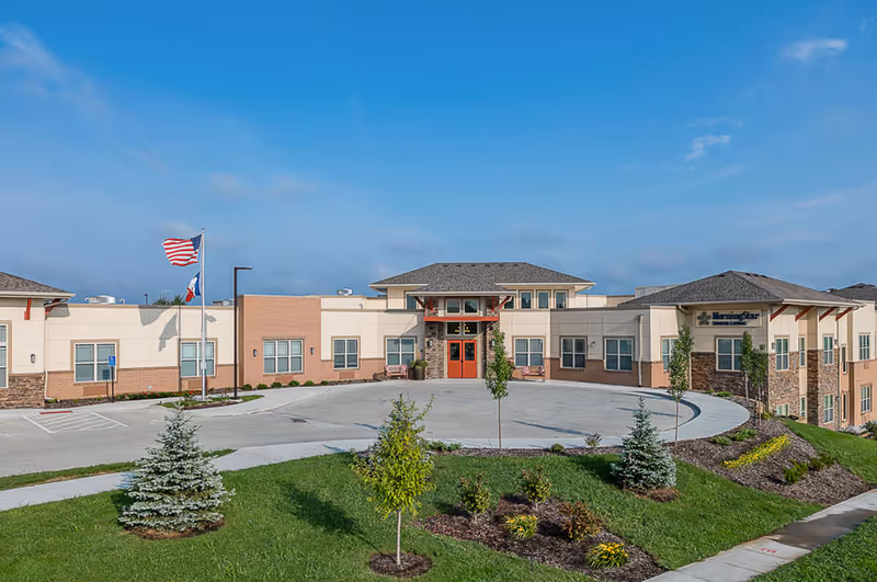 Front exterior of a single-story assisted living facility with a circular driveway, flags, and landscaped lawn under a blue sky.