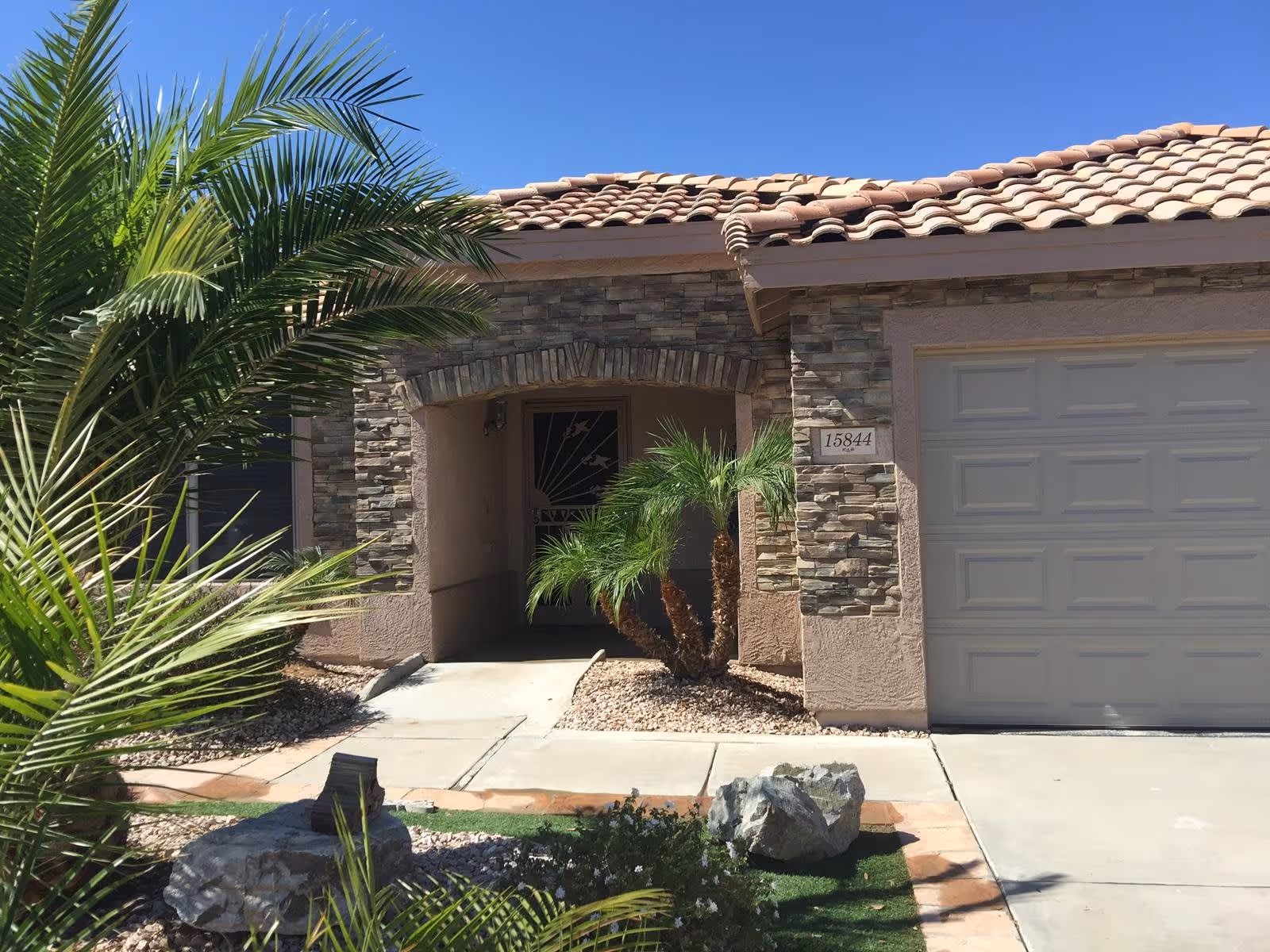 Front exterior view of a single-story house with a tiled roof, stone and stucco facade, a garage door on the right, a small covered entrance with a door, and landscaping including palm trees and rocks.