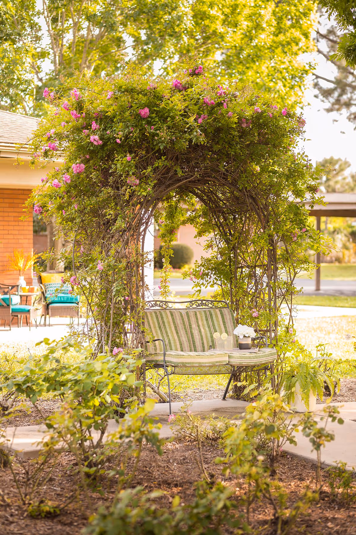A garden seating area with a metal bench that has green and white striped cushions, placed under an archway covered with green vines and pink flowers. The background shows part of a brick building, outdoor chairs, and trees with sunlight filtering through.
