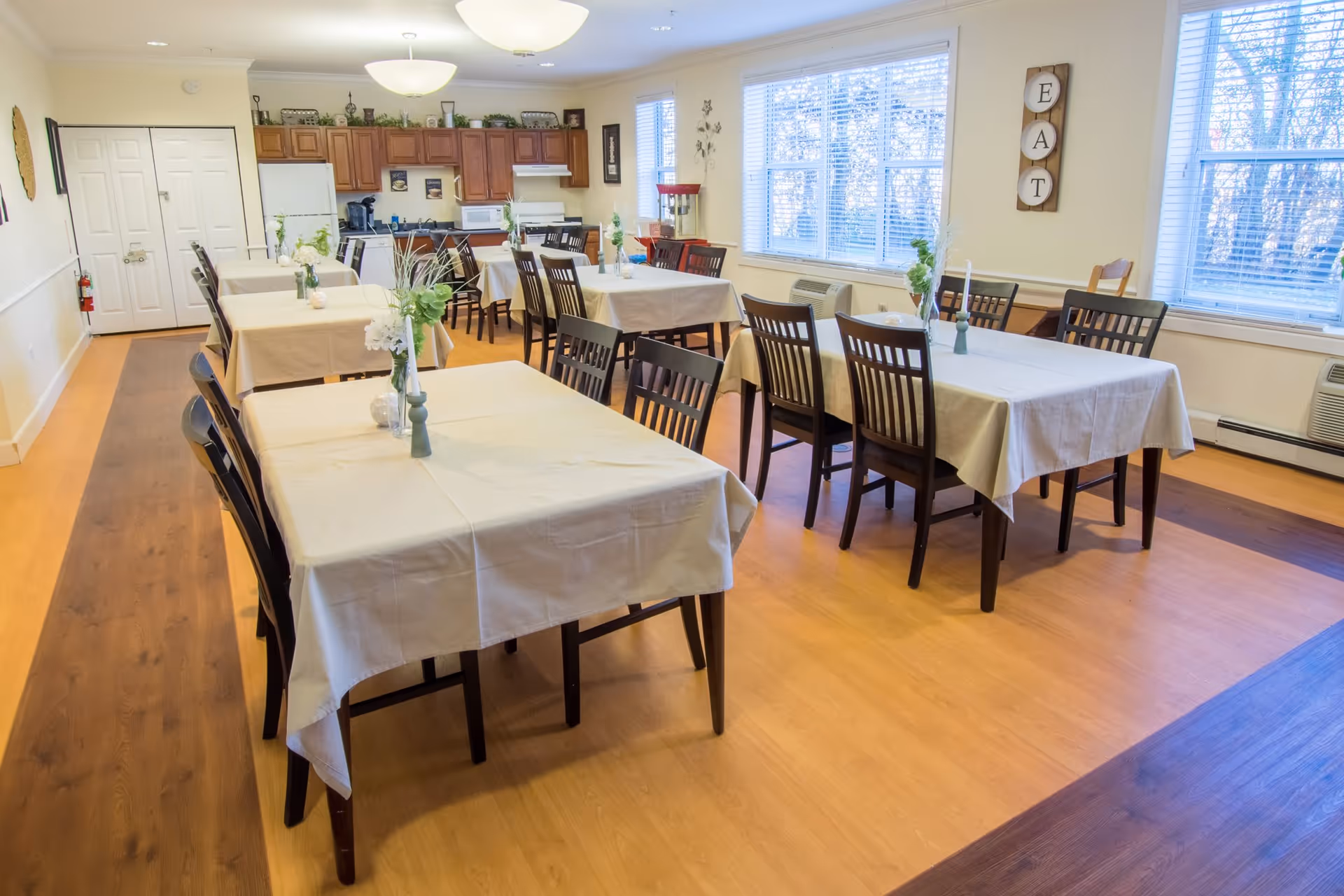 A bright dining room with several tables covered in white tablecloths, each adorned with small flower vases. The room has wooden chairs, large windows letting in natural light, and a kitchen area with wooden cabinets and appliances in the background. A wall decoration spells out the word 'EAT'.