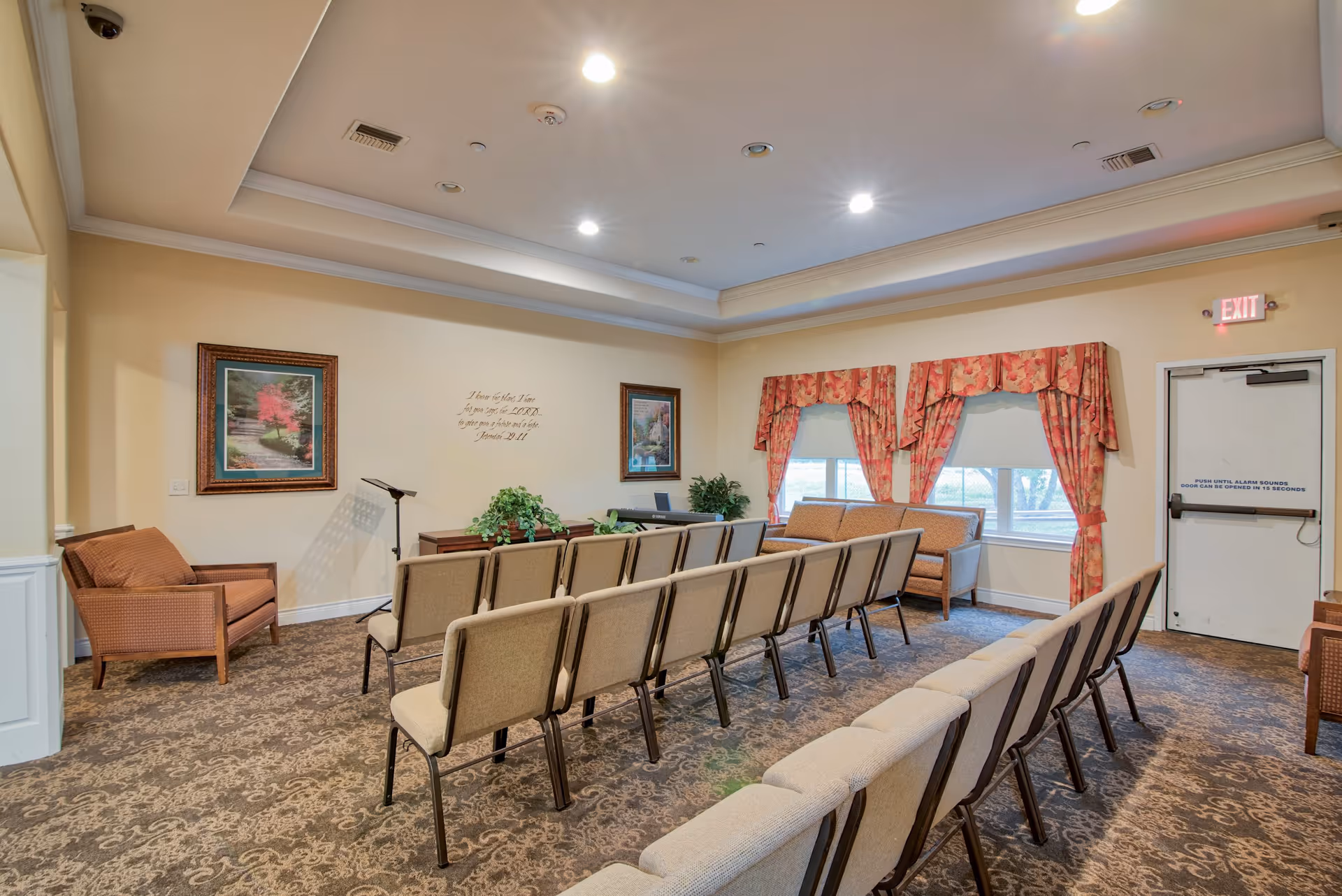 A small meeting or chapel room with rows of beige chairs facing a piano and music stand. The room has patterned carpet, floral curtains on the windows, framed artwork on the walls, and an exit door with a sign. There are also a couple of armchairs and some green plants.
