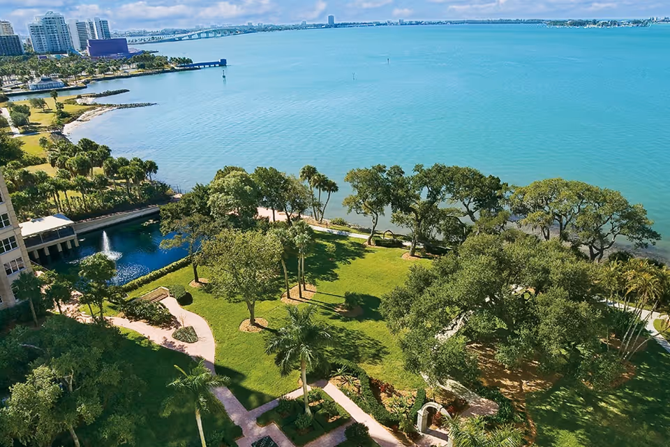 Aerial view of a waterfront park area with green lawns, trees, walking paths, and a small fountain pond next to a building. The scene overlooks a large body of blue water with a distant city skyline under a partly cloudy sky.