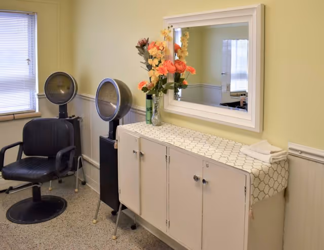 Interior view of a salon area in a senior living facility with two black salon chairs and hair dryers, a white cabinet with a patterned cloth on top, a vase with colorful flowers, a mirror on the wall, and a window with blinds.