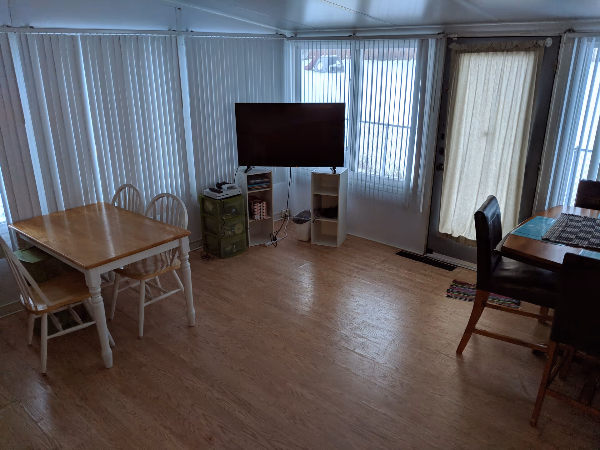 Interior view of a senior care home common area with wooden flooring, a wooden table with four chairs on the left, a flat-screen TV mounted on two white shelves in the center, and a dining table with dark chairs on the right. Vertical blinds cover the windows and a door with a curtain is visible.