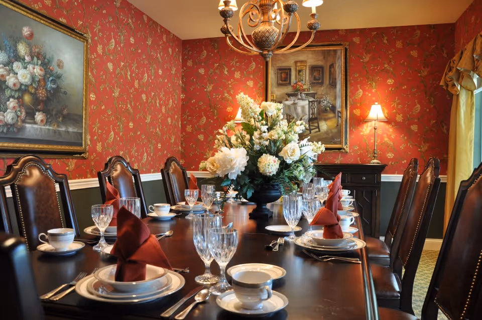 A formal dining room with a long dark wooden table set with white china, crystal glasses, silverware, and red folded napkins. The room has red floral wallpaper, two framed paintings on the walls, a chandelier overhead, and a large floral centerpiece on the table. There are leather chairs around the table and a window with yellow curtains on the right side.