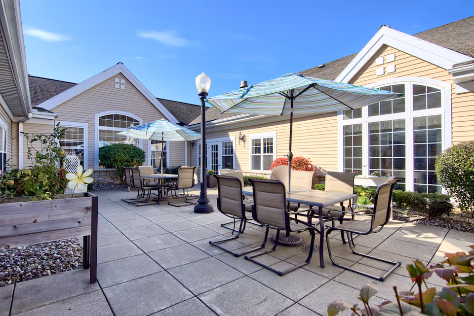 Outdoor patio area at a senior living facility with multiple tables and chairs under large striped umbrellas, surrounded by beige buildings with large windows and some greenery.