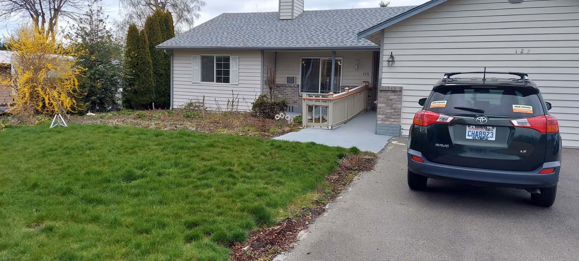 Front exterior view of a single-story house with light gray siding, a small porch with a ramp, and a driveway with a black Toyota RAV4 parked. The lawn is green with some bushes and a yellow flowering tree on the left side.