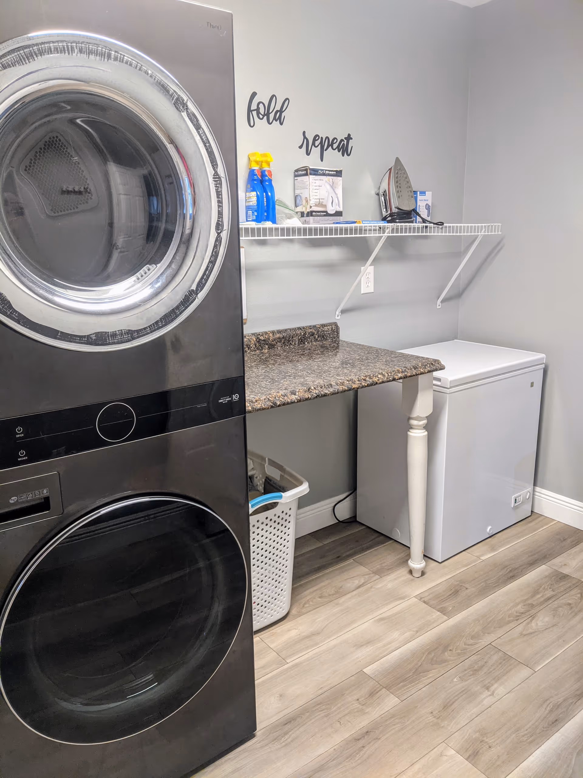 Laundry room with a stacked black washer and dryer on the left, a white laundry basket underneath a brown countertop, a white chest freezer on the right, and a white wall shelf above holding cleaning supplies and an iron. The wall has the words 'fold' and 'repeat' written above the shelf.